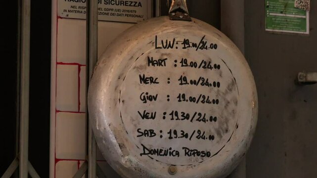 Close-up of a handmade restaurant schedule written on an old metal pot in Genoa, Italy, showing weekly opening times with unique character and vintage flair