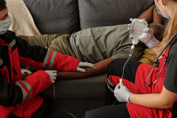 Paramedics providing medical assistance to patient lying on couch, administering IV drip and monitoring patient's state. Patient receiving proper care from trained professionals