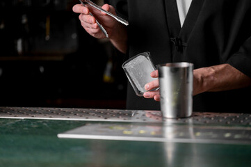 A bartender expertly prepares a cocktail, holding ice in a glass while using tongs. The scene is set in a stylish bar with a blurred background.