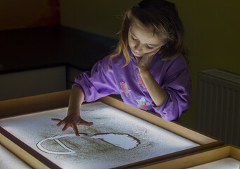 Beautiful little girl draws with sand on a light square, art therapy, sand animation, creative...