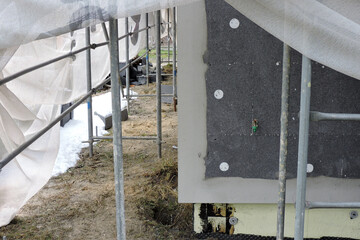 A wall of a house covered with EPS graphite polystyrene boards for thermal insulation, a scaffolding and building safety scaffolding net, some adhesive, anchors, an exterior wall mounted water faucet