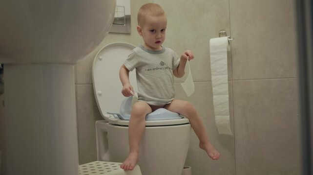 baby toddler sitting on toilet learning to clean his hygiene bottom butt anus toilet paper. potty training child in bathroom. little boy kid pulls toilet paper sweep his back after poop poo defecate