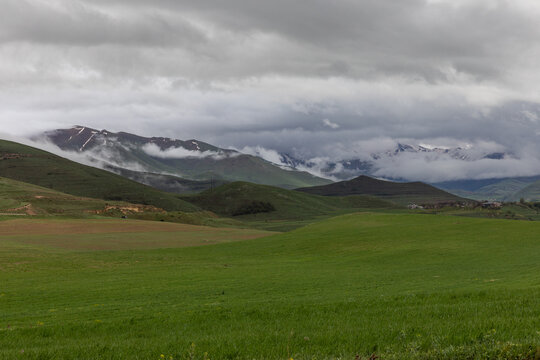 A dramatic cloudy landscape near Sisian, Armenia, showcasing lush green meadows, a calm lake, and distant snow-covered peaks partially covered by mist