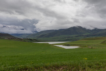 A dramatic cloudy landscape near Sisian, Armenia, showcasing lush green meadows, a calm lake, and distant snow-covered peaks partially covered by mist