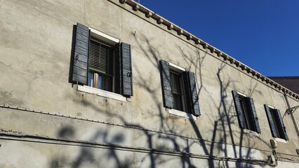An old building with a weathered facade features dark wooden shutters on its windows. Shadows of leafless tree branches stretch across the wall.