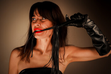 A striking woman with long dark hair bites a piece of a black leather whip, wearing elbow-length...