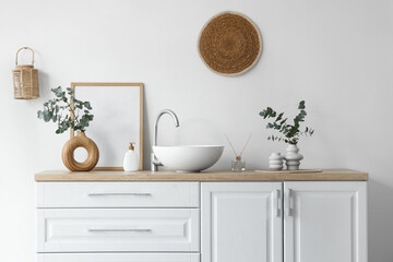 Chest of drawers with sink, photo frame and vases with eucalyptus twigs near white wall in bathroom