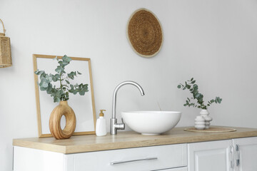 Chest of drawers with sink, photo frame and vases with eucalyptus twigs near white wall in bathroom