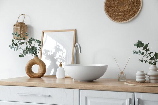 Chest of drawers with sink, photo frame and vases with eucalyptus twigs near white wall in bathroom