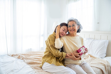 Happy Asian family adult daughter give present gift box to senior mother on bed in bedroom. Elderly...