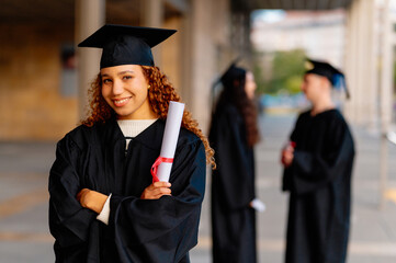 Young graduate smiling and holding diploma with other blurred graduates in the background on...