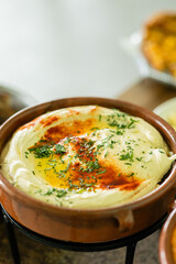 closeup of brown ceramic dish filled with creamy hummus garnished with fresh herbs smoked paprika and olive oil