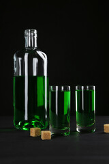 Absinthe in shot glasses, bottle and brown sugar on table against black background