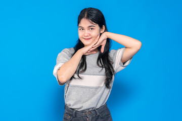 Young Asian woman posing playfully with a charming smile, resting her face on her hands. She exudes happiness and confidence against a vibrant blue background.