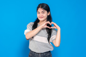 Young Asian woman smiling while forming a heart shape with her hands. She has long black hair, wears a casual gray t-shirt, and stands against a solid blue background, expressing love.
