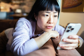 Concerned Asian woman looking at smartphone on living room couch