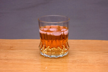 A clear glass filled with an amber-colored liquid and ice cubes is positioned on a light wooden table against a neutral gray background.