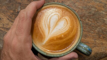 Hands clutching a cup of golden flat white coffee with love heart shaped latte art on rustic wooden table