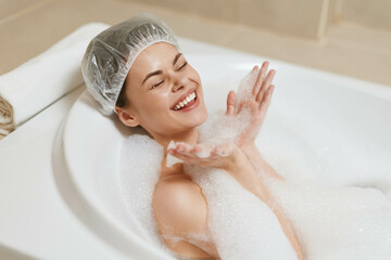 Joyful woman enjoying a relaxing bubble bath, wearing a shower cap, with soap bubbles and a serene bathroom setting. Self-care and wellness concept.