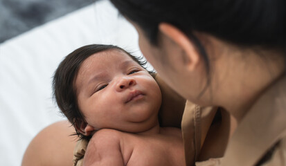 Cute multiracial newborn baby lying in arm and looking at Asian mother at home. Mom hug and comfort her little infant. Family and child care concept