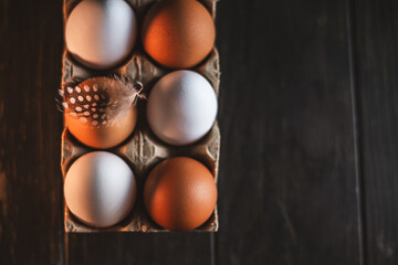 White and brown chicken eggs are nestled in a cardboard carton, one adorned with a delicate spotted feather, resting on a rustic wooden table, creating a charming still life, top view, with copy space