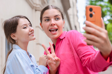 Smiling young women taking a selfie outdoors, smiling and making playful hand gestures while posing for the camera, dressed casually, capturing a fun moment in a bright and lively setting.