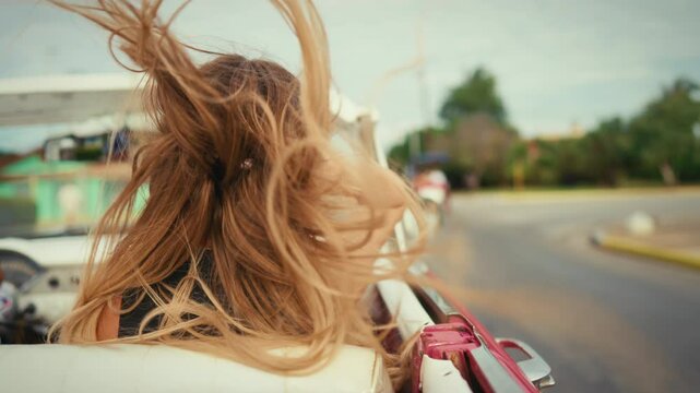 Swaying blonde hair by wind of carefree woman passenger of cabriolet in summer. Happy people traveling by car in summertime, back view of lady with long hair, slow motion, cinematic shot, weekend trip
