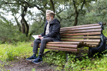Fototapeta premium Senior man writing on notepad while sitting on bench in park