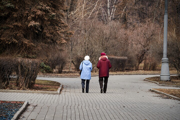Two older women walking on a brick path in a park. One is wearing a blue coat and the other is wearing a red coat
