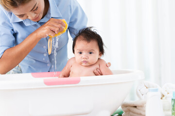 Naklejka premium Asian mother bathing her 3 months newborn baby in bathtub at home, mom cleaning her cute little daughter with liquid soap and sponge. Baby have fun bathing with mom
