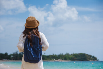 Rear view of a female traveler with hat and backpack standing on the beach by the sea
