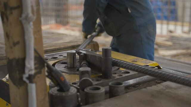 Worker in dark uniform uses bending machine to shape steel rebar. Industrial equipment ensuring precise reinforcement bar modification at site