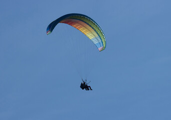 A paraglider soaring against a clear, solid blue sky. The canopy is a vibrant mix of colors, with streaks of yellow, orange, pink, blue, and green, creating a striking visual contrast.