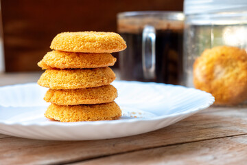 Stack of golden coconut cookies on a white plate, shallow depth of field. Coffee and a jar of cookies blurred in the background. Rustic wooden table.