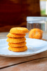 Stack of five golden cakes coconut cookies on white plate. Blurred coffee mug and jar in background. Casual food setting. Natural light. Homemade treat.
