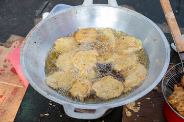 Bubbling oil fries tempeh at an outdoor street food cart. Cardboard and a dark surface surround the silver pan