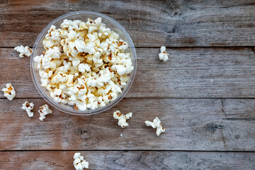 Top-down view of popcorn in clear bowl on rustic wood table. Scattered kernels on textured surface. Simple snack, neutral background.