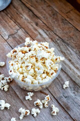 Close-up of fluffy white popcorn in a clear bowl on a weathered wooden table. Scattered kernels add casual charm. Natural light highlights the texture.