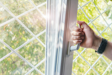Male hand opens a plastic window with a metal grate, letting in fresh air and sunlight. Manual opening white plastic pvc window at home.