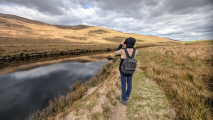 Beautiful woman backpacker in hooded jacket hiking at river Erriff near Aesleagh, county Mayo, Ireland, Irish landscape, mountains and nature background	