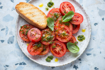 Sliced red tomatoes with basil oil, fresh basil and ciabatta on a grey plate, horizontal shot on a white and blue granite background