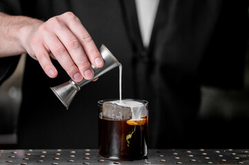 A bartender skillfully pours cream into a cocktail glass containing ice and a slice of orange,...