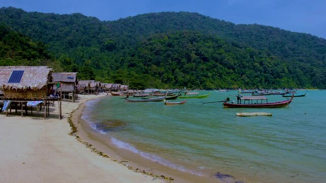 Experience the serene charm of Surin Island Thailands coastline, where traditional fishing boats gently sway in the turquoise waters. Discover lush green hills, sandy shores, and vibrant local culture