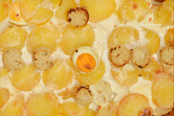 A top-down shot of a baked dish made with cooked potato slices, sausage slices, hard-boiled egg slices, and a creamy sauce with egg, showcasing a golden brown crust