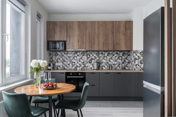 built-in kitchen with grey and wooden cabinets, wooden countertop, backsplash. modern furniture. microwave, steel refrigerator. white peonies in vase, fruits, berries, glasses on wooden kitchen table