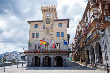 main facade of the town hall of Castrourdiales, with the flags waving in the wind, with its empty square of people, and its typical arcaded arcades, on a cloudy summer day