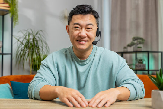 Asian Chinese man wearing headset, freelance worker call center or support service operator helpline. Chinese mature guy having talk with client or colleague communication support waving hello at home