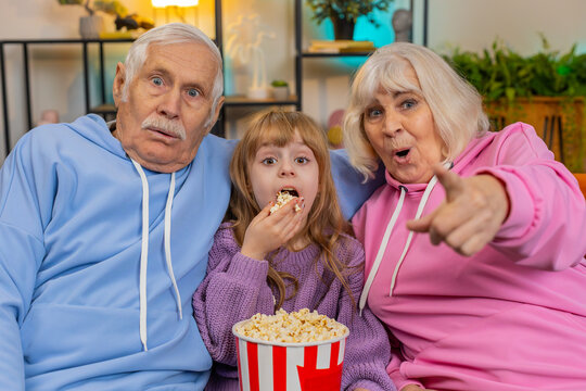 Happy Caucasian grandfather, grandmother and granddaughter eating popcorn and watching movie on sofa at home. Smiling small girl with grandparents enjoying film during weekend in living room apartment