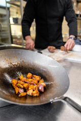 Close-up of sautéed vegetables in a frying pan, with a chef in the background preparing other ingredients in a restaurant kitchen