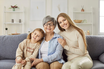 Portrait of happy smiling family of mother, grandmother and child girl hugging while resting on couch at home and looking cheerful at camera. Love, generations and Mothers day concept.
